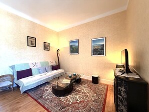 A furnished living room with a sofa, a coffee table, a television, and framed pictures on the wall. - T3 Apartment mountain view - 5 guests (La Bourboule)
