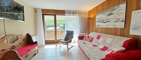 Une pièce avec un canapé rouge, une chaise en bois, un lit avec une literie à motifs, une télévision et un balcon avec vue.