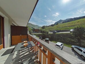 A balcony with a table and chairs, overlooking a road with cars and a mountainous landscape. - Studio cabin with slope view - 4 guests (Le Grand-Bornand)