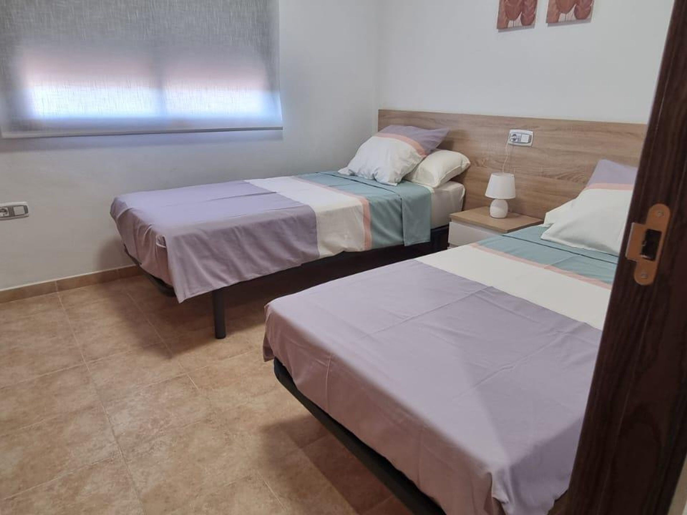 Een kamer met twee eenpersoonsbedden, een nachtkastje met een lamp, een raam met zonwering en een stopcontact.