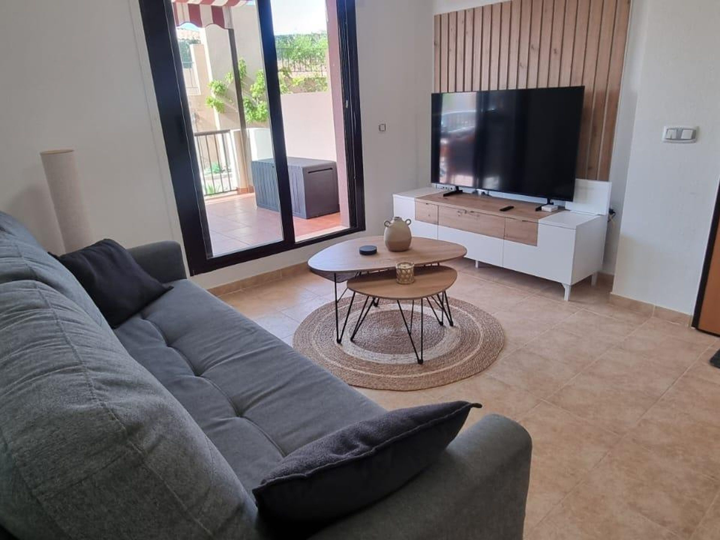 A modern living room with a grey sofa, a coffee table, a flat-screen TV on a wooden cabinet, and a sliding glass door leading to an outdoor area.