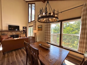 A cozy living room with a fireplace, a television, a dining table with chairs, and large windows offering a view of greenery. - Updated 3 bedroom condo- walking distance to ski slopes! (Sugar Mountain)
