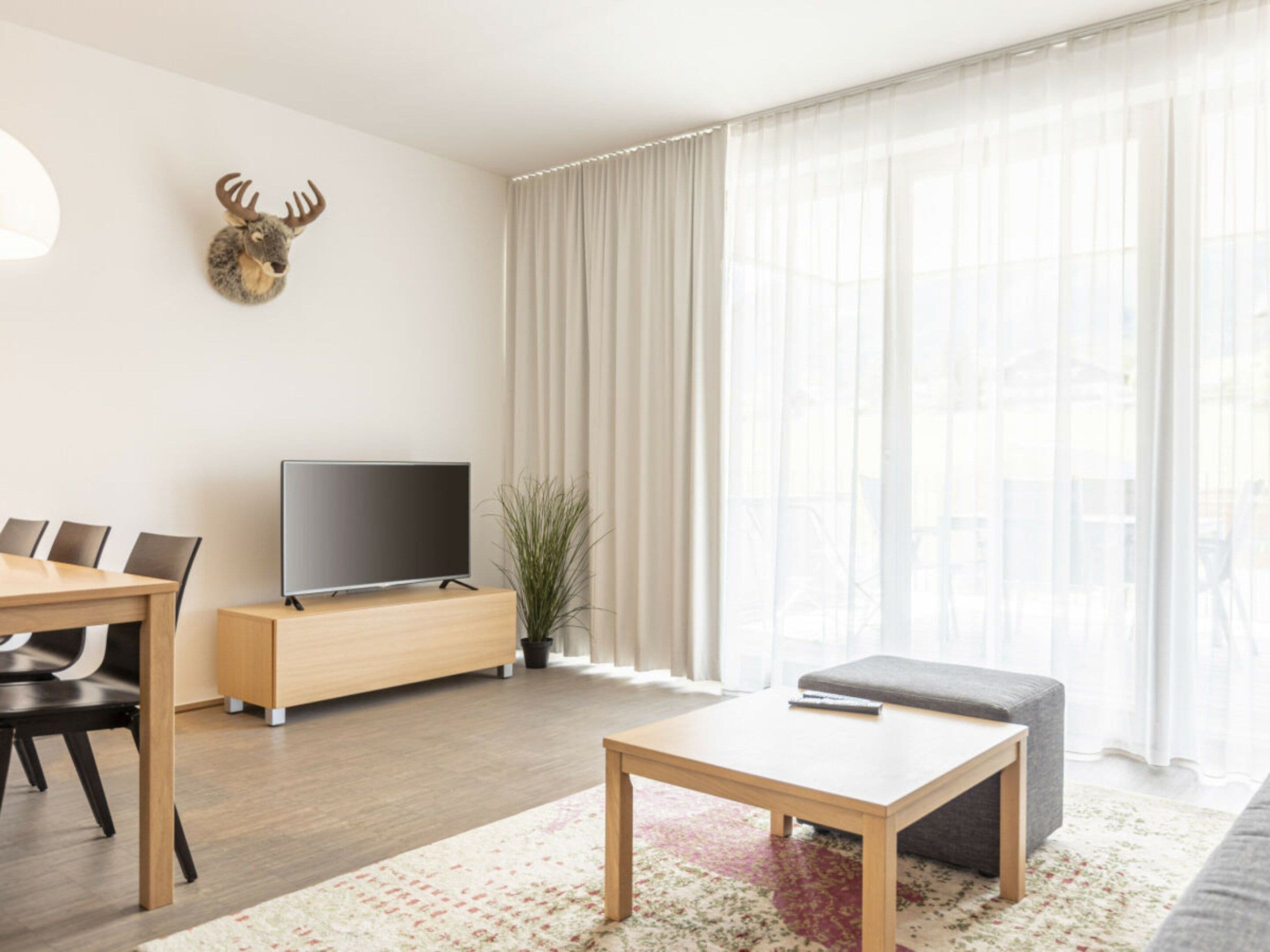 A modern living room with a flat-screen TV, a dining table with chairs, a sofa, and a decorative deer head on the wall.