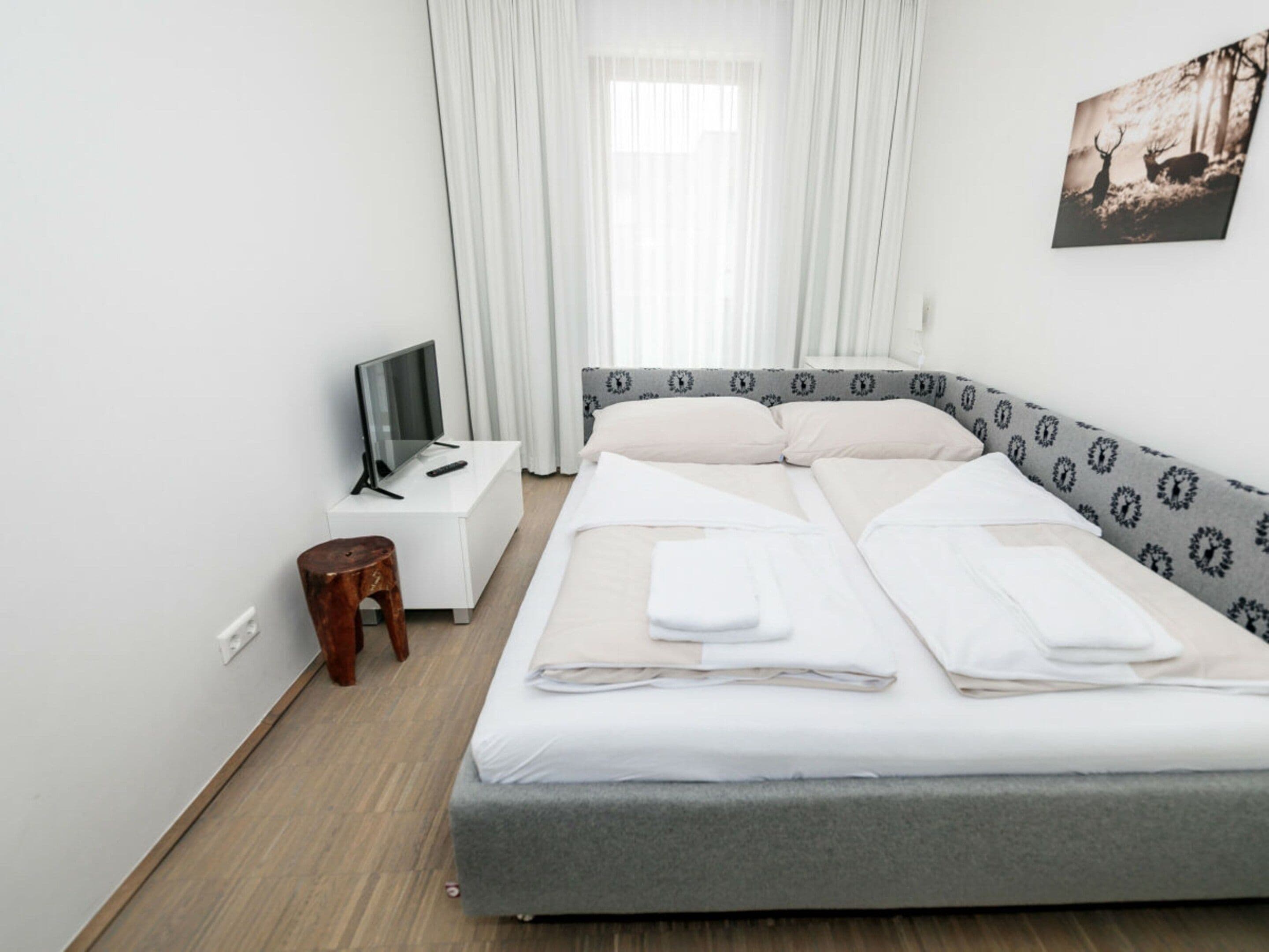 A hotel room with a bed, a TV on a nightstand, a wooden stool, and a painting on the wall.