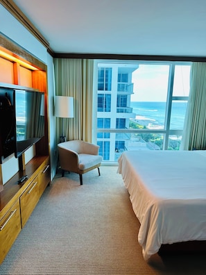 A hotel room with a bed, a TV, a chair, a desk, and a large window offering a view of the ocean and buildings. - Ultimate 2BR Oceanfront Carillon Beach Getaway (Miami Beach)
