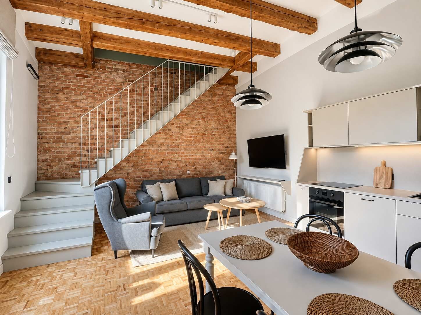 A modern living space with a brick wall, a staircase, a grey sofa, a dining table with woven placemats, and a kitchen area.