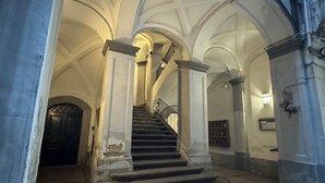 Uma imponente escadaria com teto abobadado, que leva a um espaço interior com colunas e outra escada.