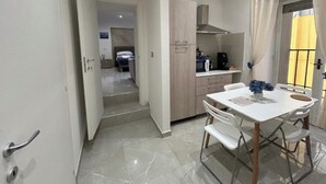 Cozinha moderna com área de jantar, destacando uma mesa e cadeiras brancas, máquina de café e vista pela janela com cortinas.