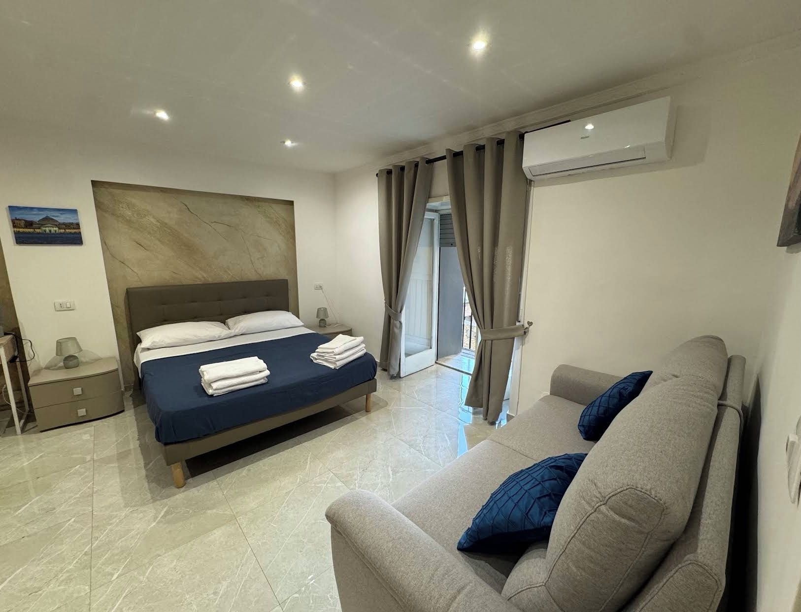 A hotel room with a bed, a sofa, an air conditioning unit, and a view through a door.