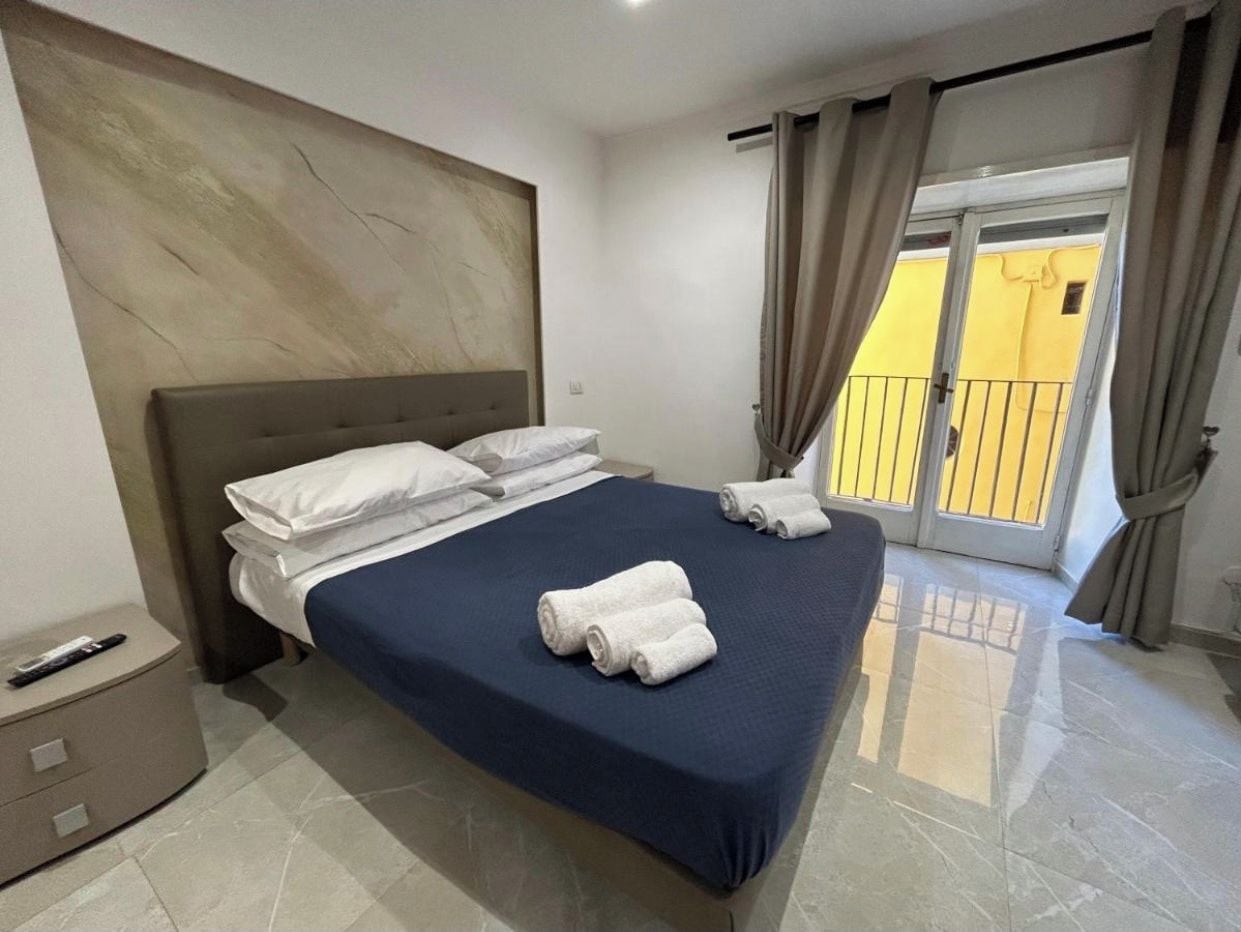A hotel room with a bed, a bedside table, a nightstand, and a view of the outside.
