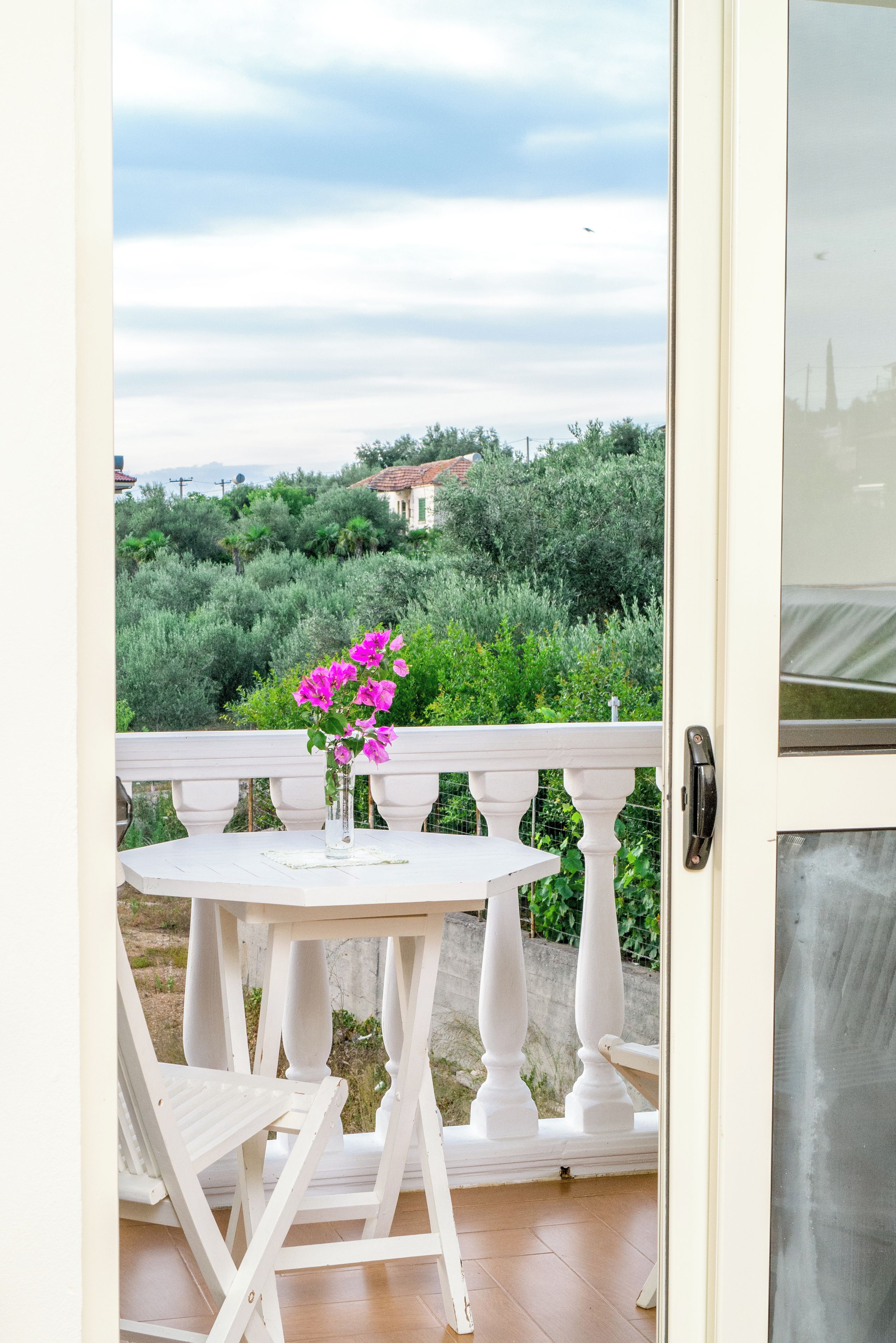 A balcony with a table and chairs, a vase with flowers, and a view of greenery and a building in the distance.