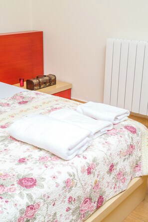 Un lit bien fait, recouvert d’une couvre-lit à motifs floraux, une table de chevet avec une lampe et un radiateur fixé au mur.