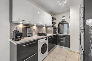 A modern kitchen with a built-in washing machine, a coffee machine, and a large stainless steel range hood. - Apartment Giono by Sur la mer (Fréjus)