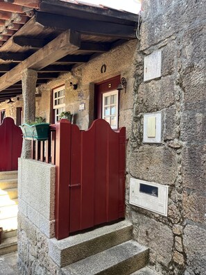 A stone building with a red gate, steps leading to a door, and a small porch area with a green chair. - Ant House (Ponte da Barca)