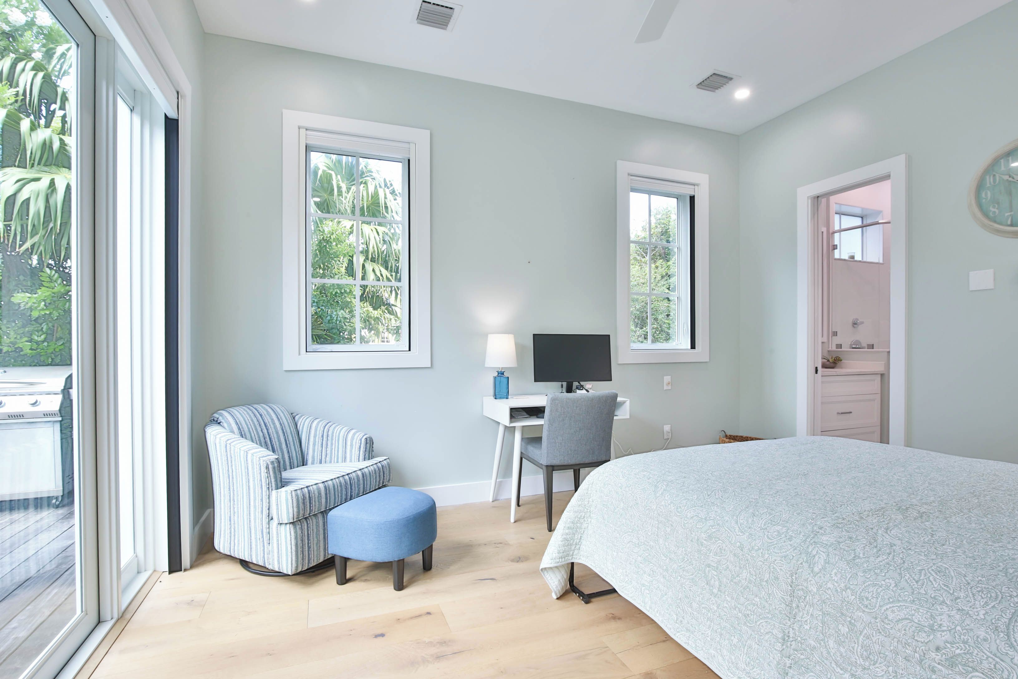 A bedroom with a bed, a desk with a computer, a chair, and a window with a view of greenery.