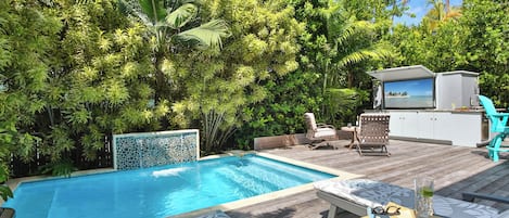 A swimming pool with a wooden deck, lounge chairs, and a bar area with a TV.