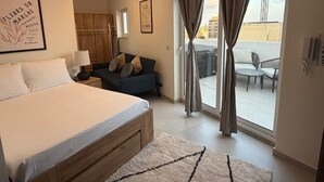 Un dormitorio con cama, sofá, ventana con cortinas, lámpara de pared y un cuadro enmarcado.