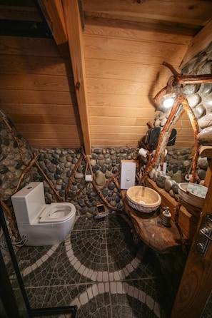 A rustic bathroom with a stone wall, wooden beams, a white toilet, and a stone basin.