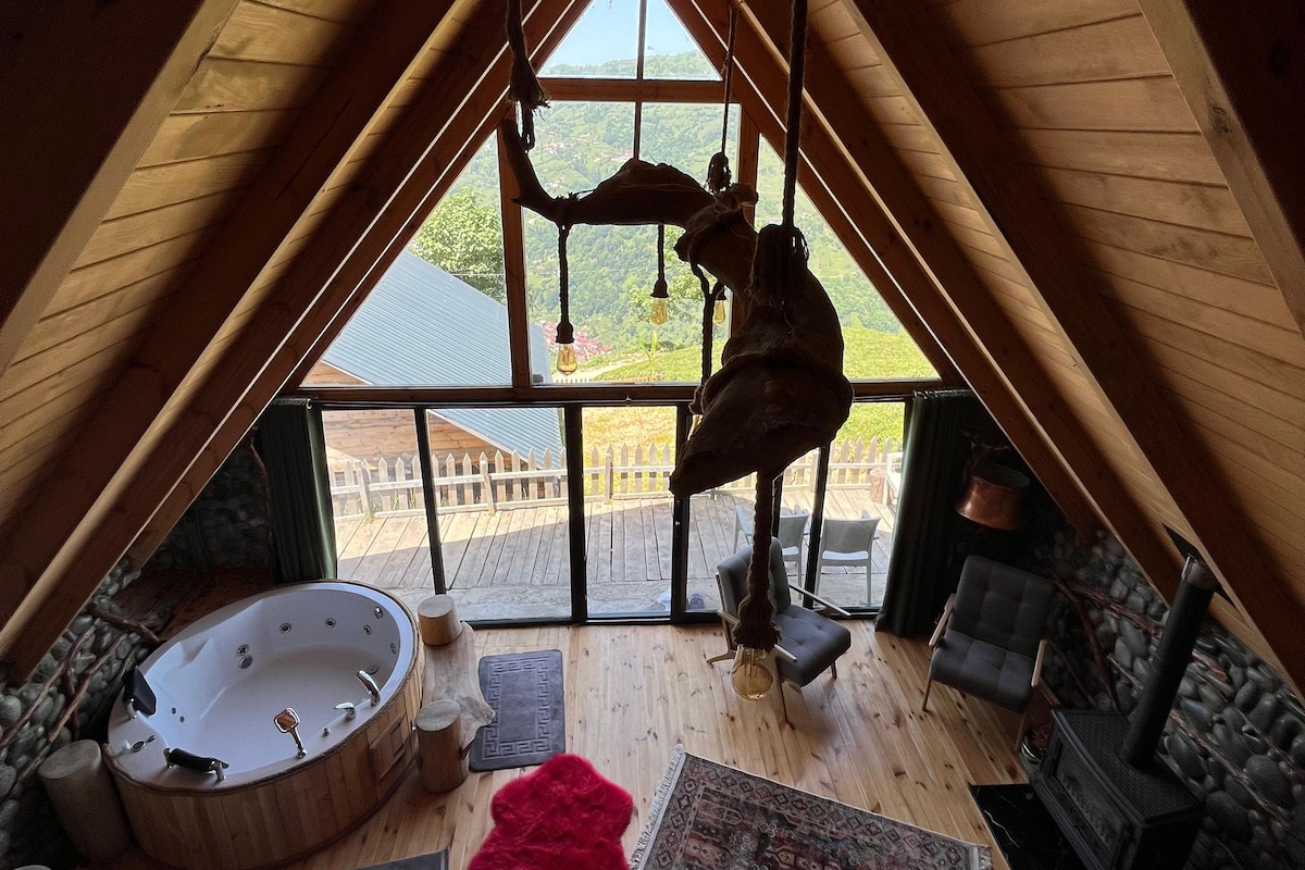 A cozy attic room with a large window, a wooden ceiling, a stone fireplace, a sofa, a red rug, and a hot tub.