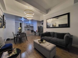 Ein modernes Wohnzimmer mit einem grauen Sofa, einem beigen Hocker und einem Essbereich mit Tisch und Stühlen.