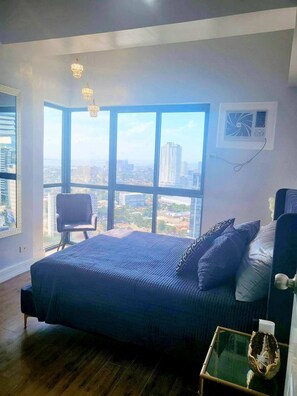 Ein Schlafzimmer mit großem Fenster, einem Bett mit blauer Bettdecke, einem Stuhl und einer Klimaanlage.