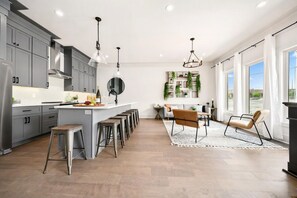 Cozinha moderna com armários cinzas, ilha central com banquetas, iluminação pendente e área de jantar com sofá e cadeiras.
