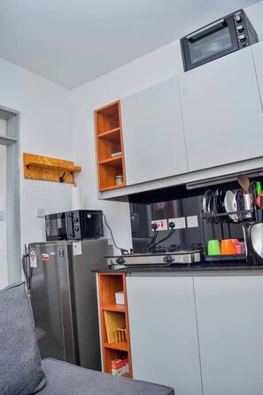 A compact kitchen with a microwave, a refrigerator, and a sink. There are wooden shelves above the counter and a small wooden cabinet below. - The Sunset Nest - Serene 1-bedroom apartment perfect for relaxing stay (Kiambu Province)