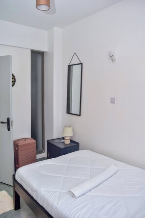 A bedroom with a bed, a nightstand, a lamp, a mirror, a door, and a suitcase. - The Sunset Nest - Serene 1-bedroom apartment perfect for relaxing stay (Kiambu Province)