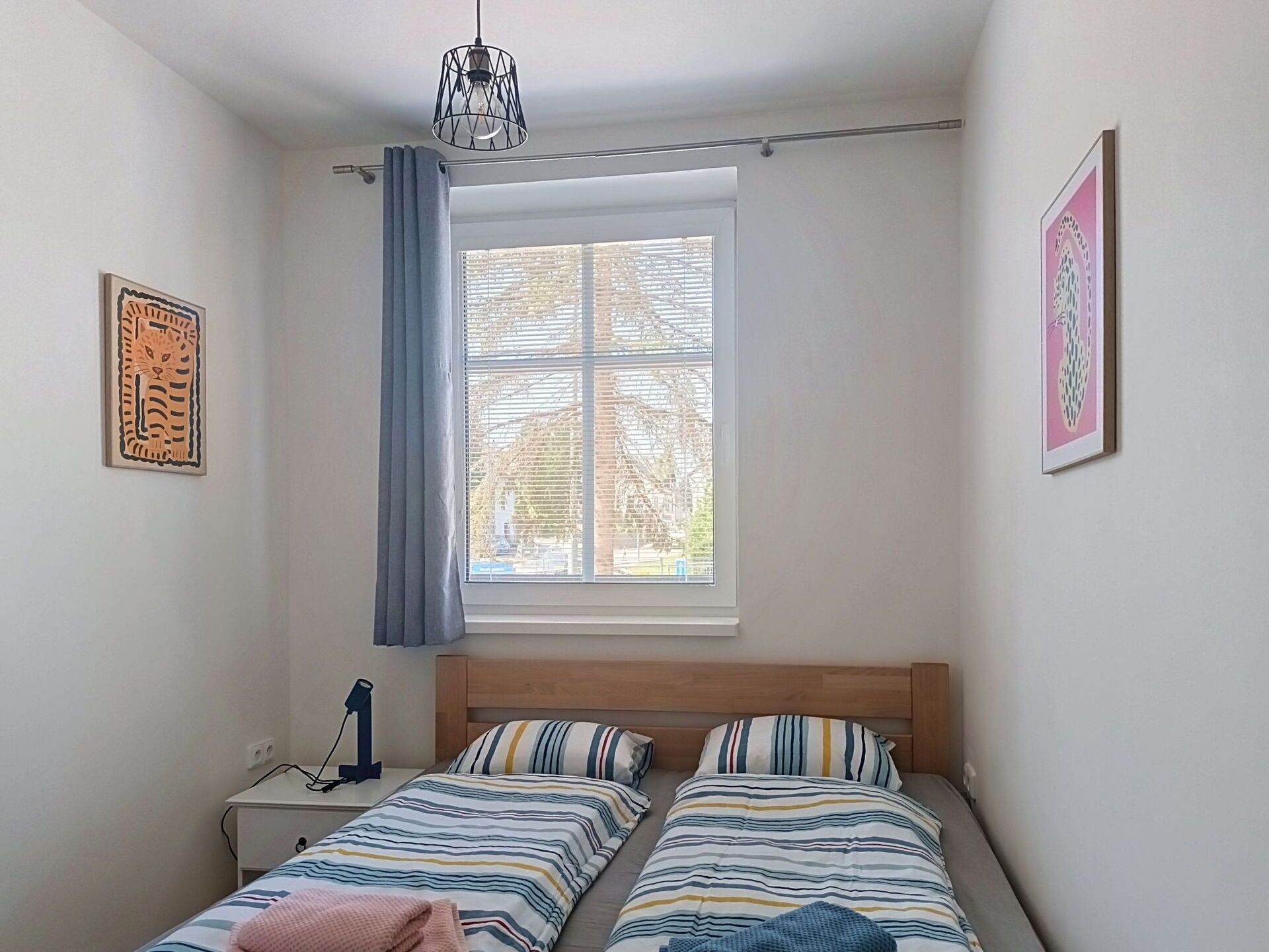 A bedroom with two beds, a window with blinds, a bedside table with a lamp, and artwork on the walls.