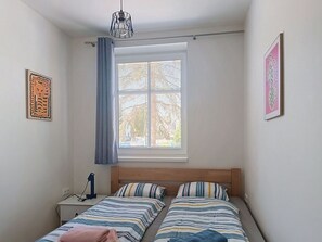 Ein Schlafzimmer mit zwei Betten, einem Fenster mit Jalousien, einem Nachttisch mit Lampe und Kunstwerken an den Wänden.