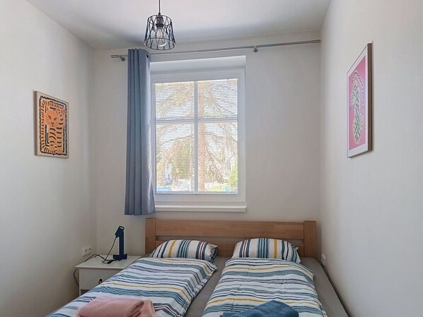 Ein Schlafzimmer mit zwei Betten, einem Fenster mit Jalousien, einem Nachttisch mit Lampe und Kunstwerken an den Wänden.