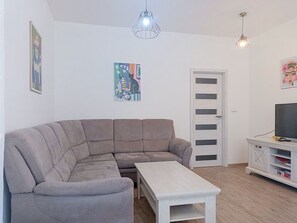 Ein Wohnzimmer mit einem grauen Ecksofa, einem weißen Couchtisch, einem Fernseher auf einem weißen TV-Schrank und einer Tür mit Fenster.