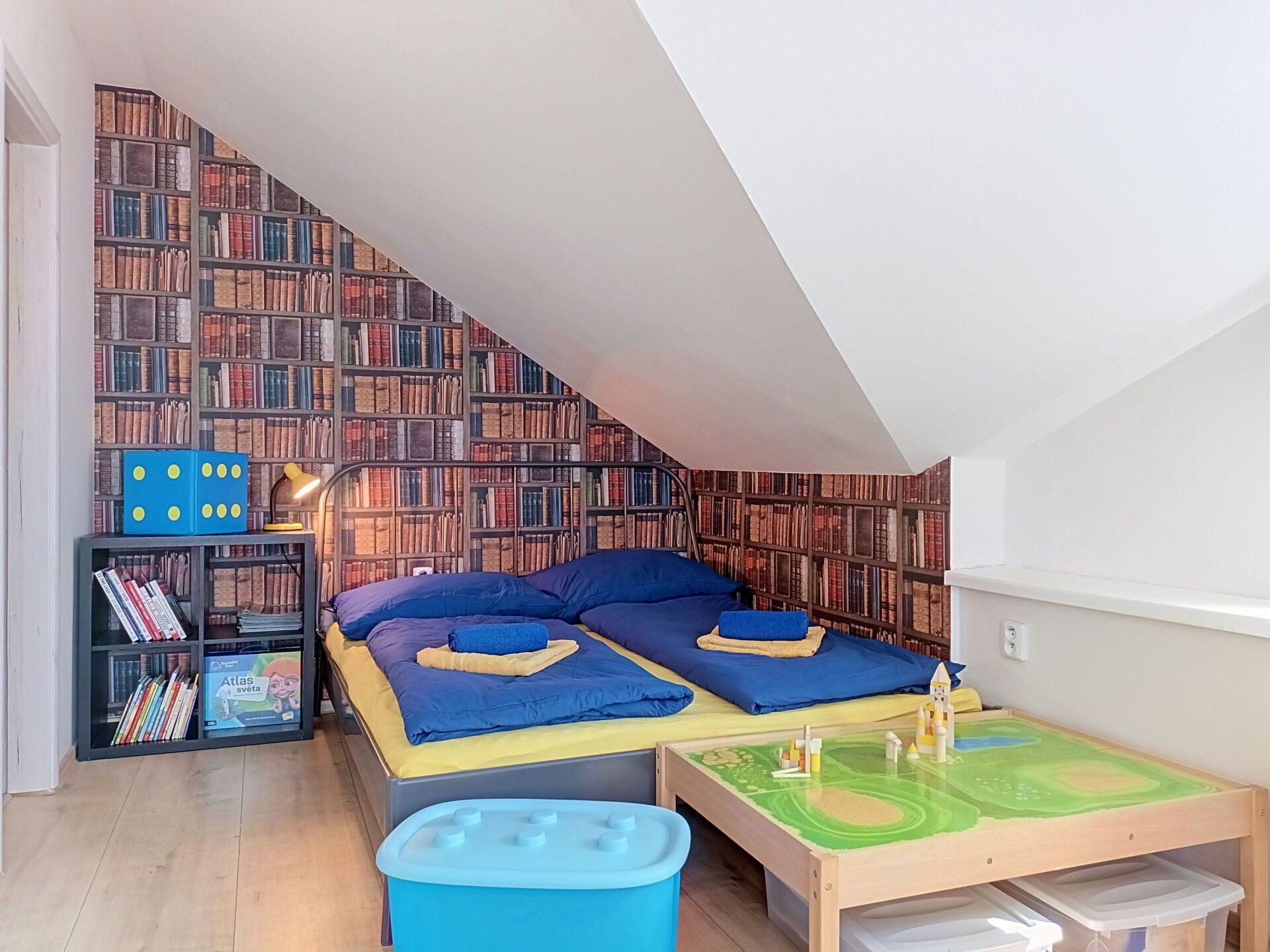 Un dormitorio con techo inclinado, una cama con ropa de cama azul, una estantería y una mesita con superficie verde.