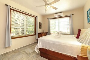 Ein Schlafzimmer mit einem Bett, einem Nachttisch, einem Deckenventilator und großen Fenstern mit Jalousien.