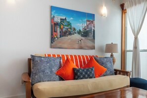 Een woonkamer met een bank, kussens, een schilderij, een lamp en een raam met gordijnen.