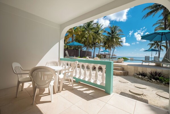 Eine sonnendurchflutete Terrasse mit Blick auf einen Strand, weiße Plastikstühle, ein Tisch und ein türkis-farbenes Geländer.