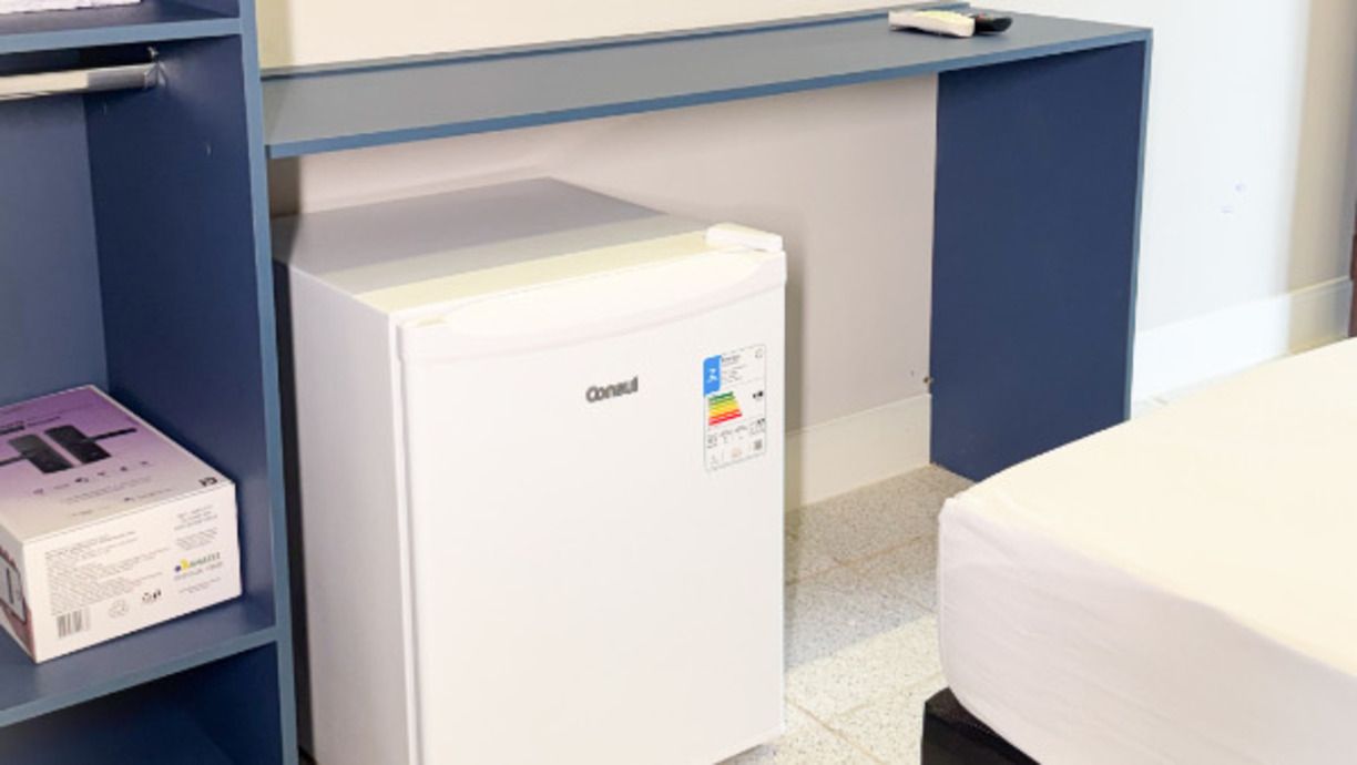 A small white refrigerator with a sticker, a white box on a shelf, and a bed with a white cover.