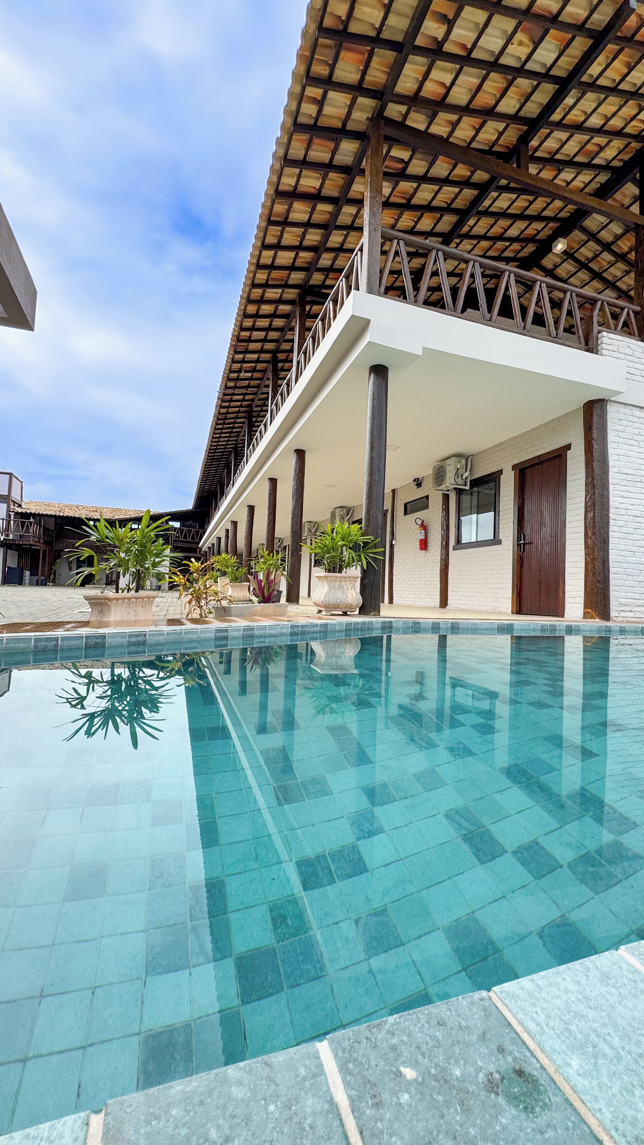 Una piscina con borde de azulejos, rodeada de plantas en macetas y una zona cubierta con columnas.