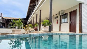 Una piscina con borde de azulejos, rodeada de plantas en macetas y una zona cubierta con columnas.