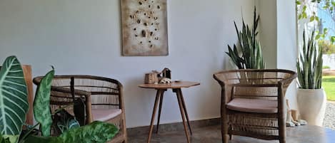 Una habitación con dos sillas de mimbre, una mesita, una obra de arte en la pared y plantas en macetas.