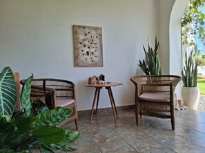 A room with two wicker chairs, a small table, a wall-mounted artwork, and potted plants.