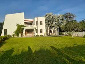 Um edifício moderno de dois andares com telhado plano, entrada central com varanda coberta e gramado bem cuidado na frente.