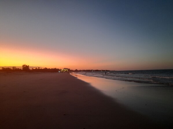 A beach scene at sunset with a calm sea, a clear sky, and distant buildings.