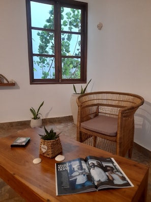 A room with a wooden table, a wicker chair, a magazine, and a window with a view of trees.