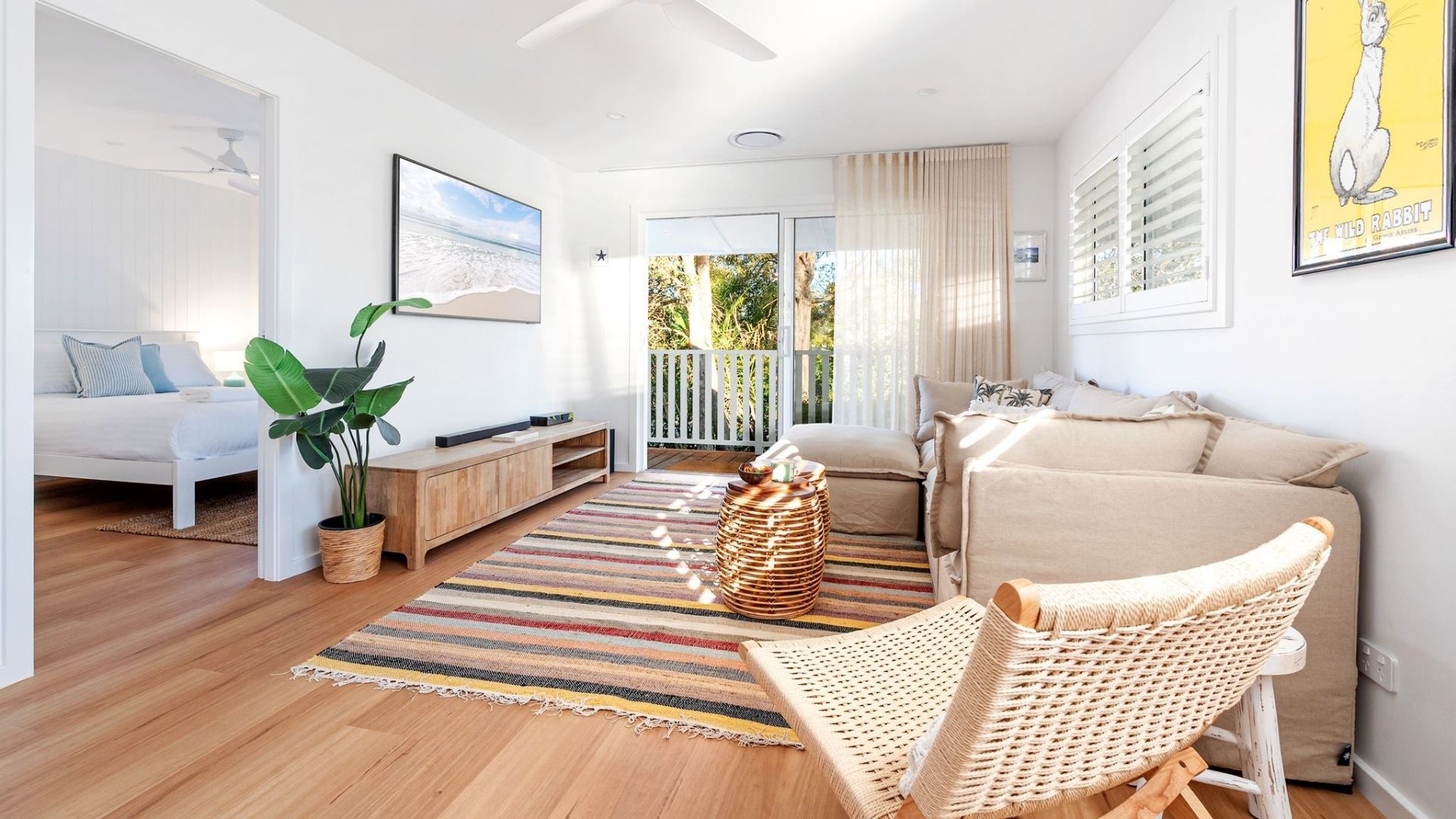 Un salon moderne avec un canapé, une table basse et un fauteuil en osier. Une grande fenêtre, ornée de rideaux, offre une vue sur la verdure environnante.