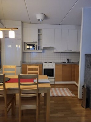 A compact kitchen with white cabinets, a stove, and a microwave. There is a dining table with chairs and a rug on the floor. - Karhunvartijan Kartano B2 | 1BR Apt in Ruka Centre (Ruka)