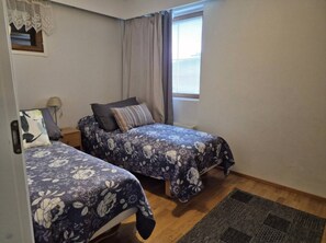 A bedroom with two beds, a window with curtains, a small bedside table with a lamp, and a rug on the floor. - Karhunvartijan Kartano B2 | 1BR Apt in Ruka Centre (Ruka)