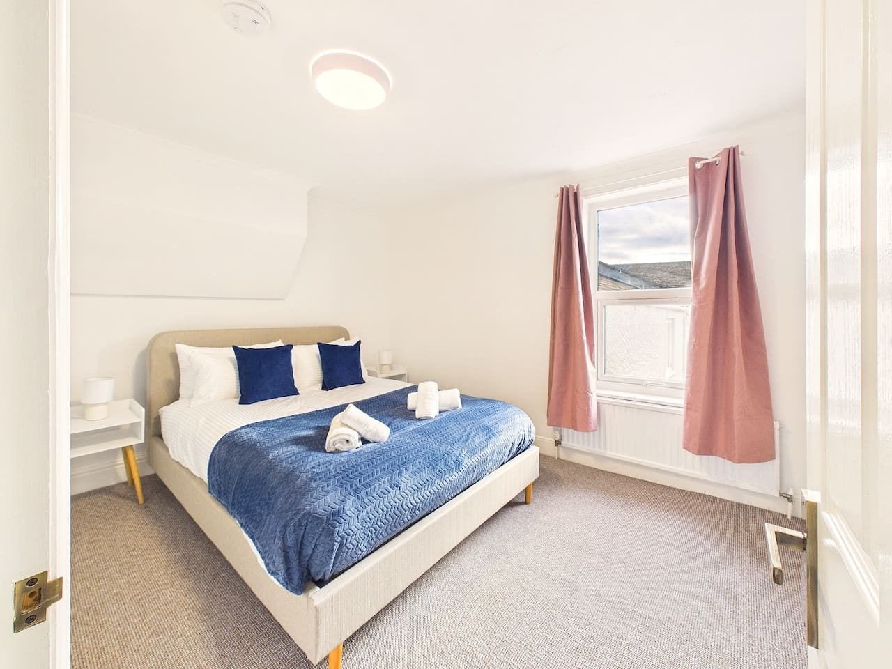 A hotel room with a bed, a nightstand, a window with curtains, and a view of the outdoors.