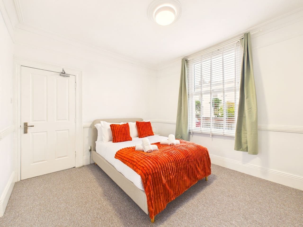 A hotel room with a bed, a window with curtains, a door, and a carpeted floor.