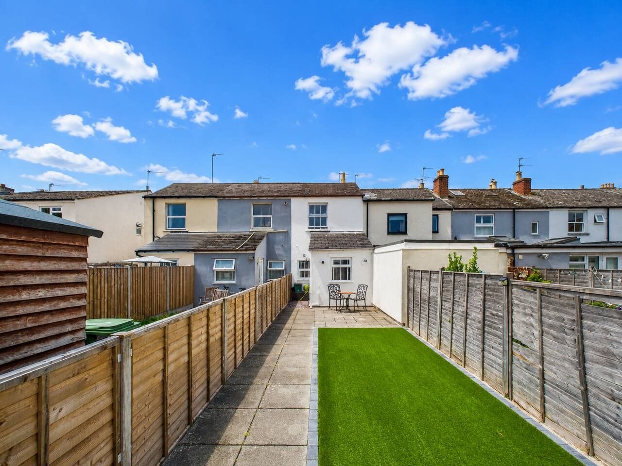 A residential area with a paved pathway leading to a green lawn, surrounded by wooden fences and houses with tiled roofs.